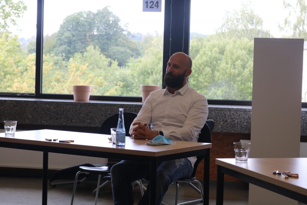 Ein Mann sitzt an einem Tisch in einem hellen Raum mit Fenstern und Pflanzen.