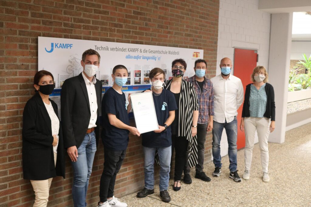 Gruppenfoto von Schülern und Lehrern mit Urkunde in der Schulumgebung. Alle tragen Masken.