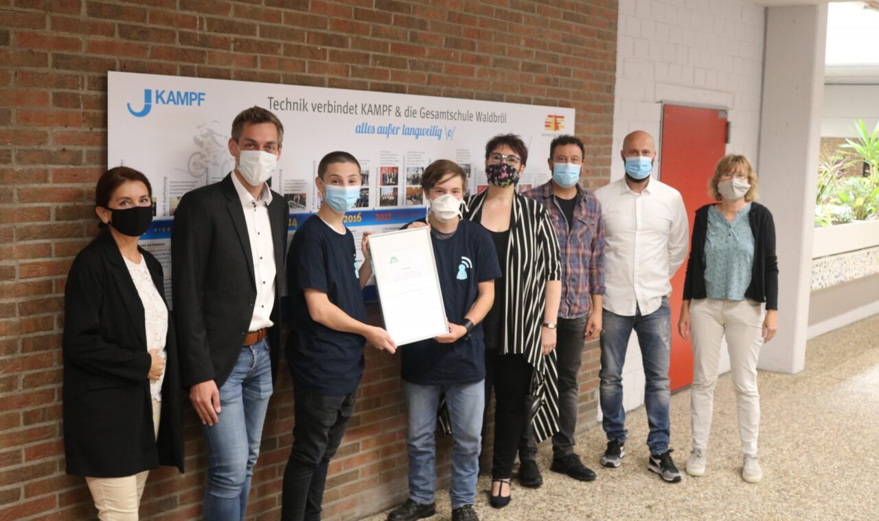 Gruppenfoto von Schülern und Lehrern mit Urkunde in der Schulumgebung. Alle tragen Masken.
