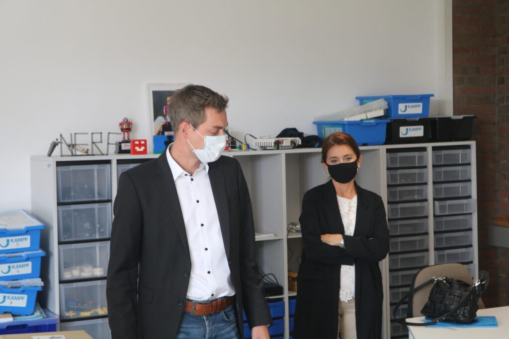 Personen in einem Klassenraum, tragen Masken, Regale im Hintergrund.