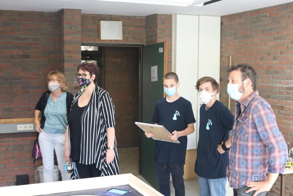 Gruppe von Personen in einem Raum, einige tragen Masken, alle schauen interessiert.