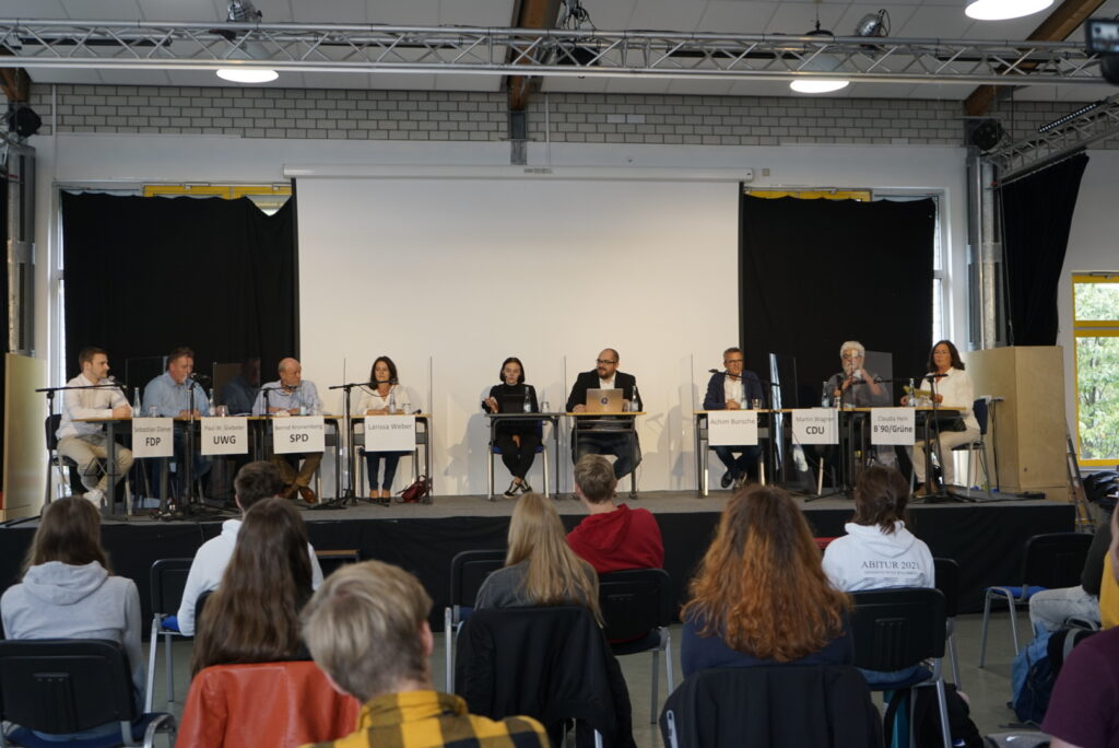 Podiumsdiskussion mit mehreren Rednern vor Publikum in einem Schulraum.