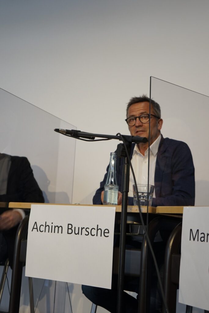 Diskussionsrunde mit zwei Personen, transparente Trennwände und Mikrofon sichtbar.