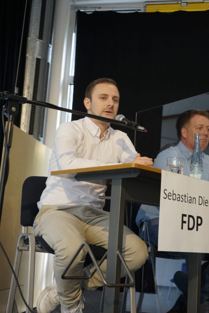 Ein junger Mann spricht an einem Tisch mit Mikrofon, FDP-Logo sichtbar.