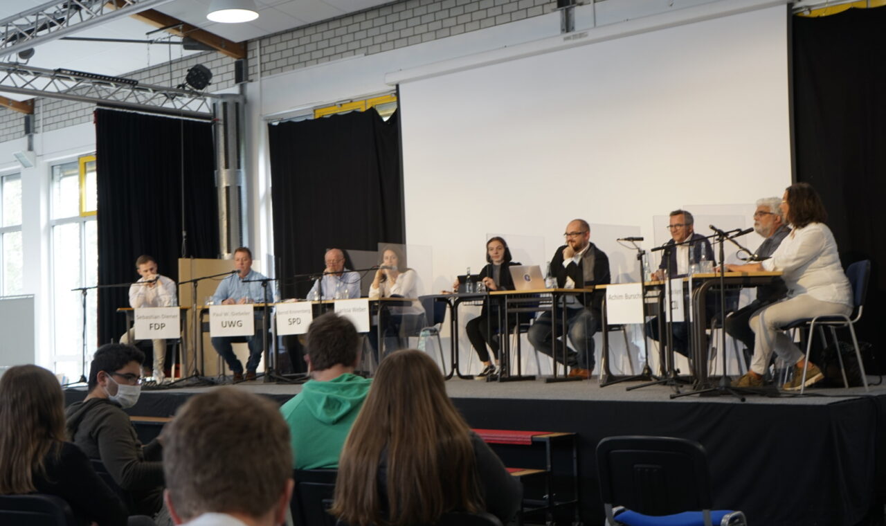 Podiumsdiskussion mit mehreren Sprechern auf der Bühne vor Publikum.