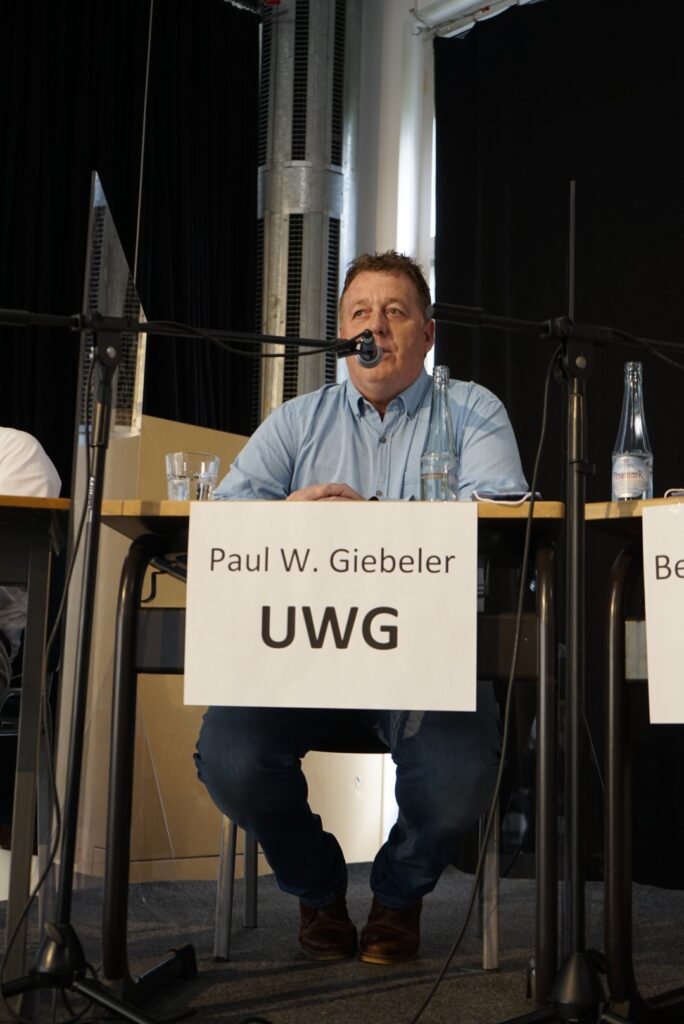 Mann spricht an einem Tisch während einer Diskussion, vor ihm ein Schild mit "UWG".
