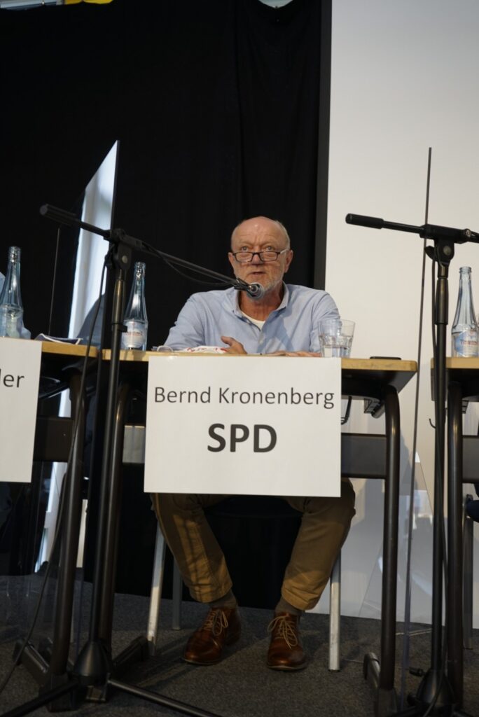 Mann sitzt an einem Tisch mit Mikrofon und Flasche, beschriftet mit „Bernd Kronenberg SPD“.