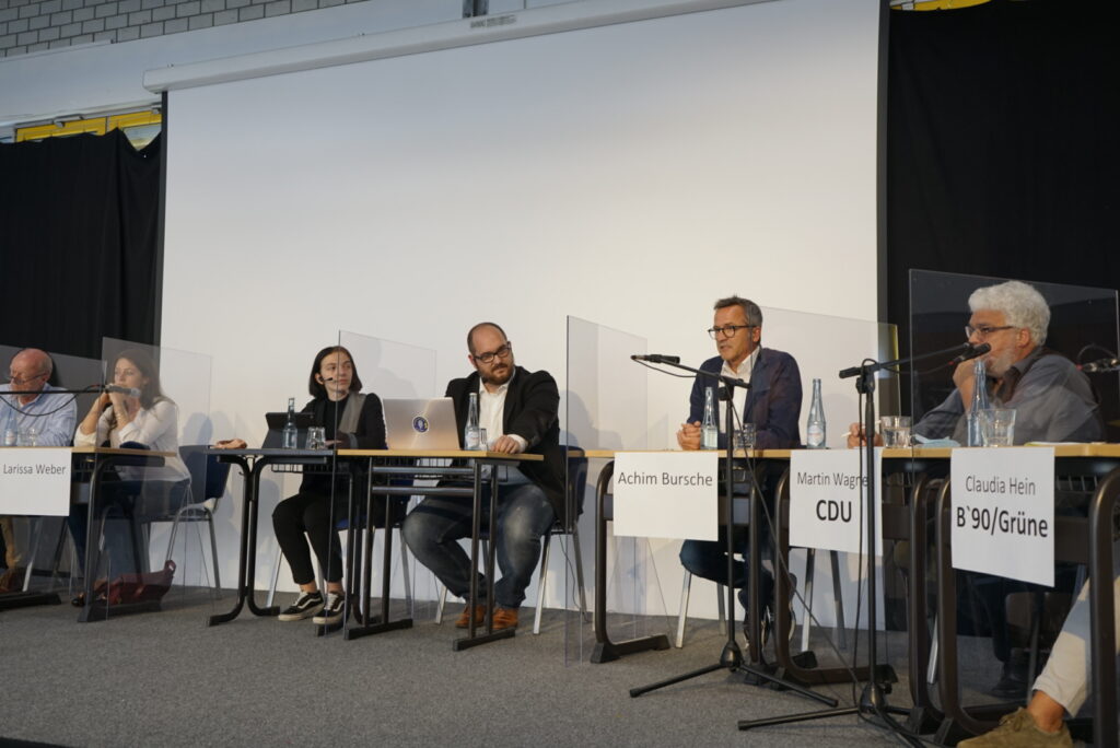 Podiumsdiskussion mit mehreren Sprechern und Mikrofonen auf einer Bühne.