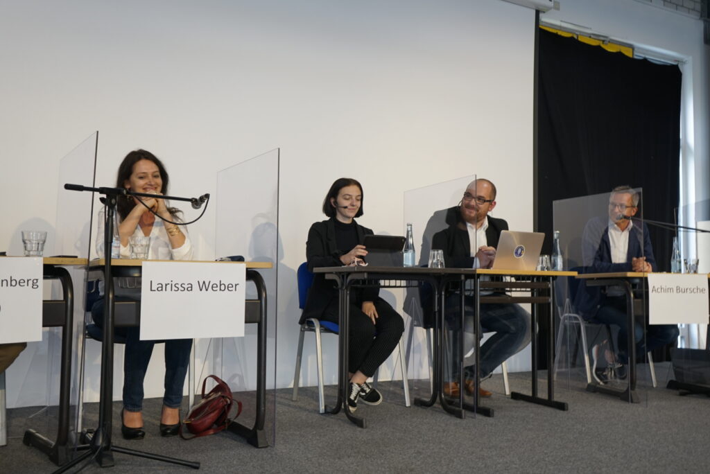 Diskussionsrunde mit vier Teilnehmern an Tischen, Mikrofone und transparente Trennwände.