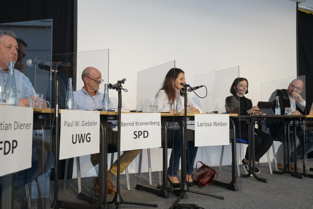 Podiumsdiskussion mit fünf Rednern und Mikrofonen, trennende Plexiglasscheiben.