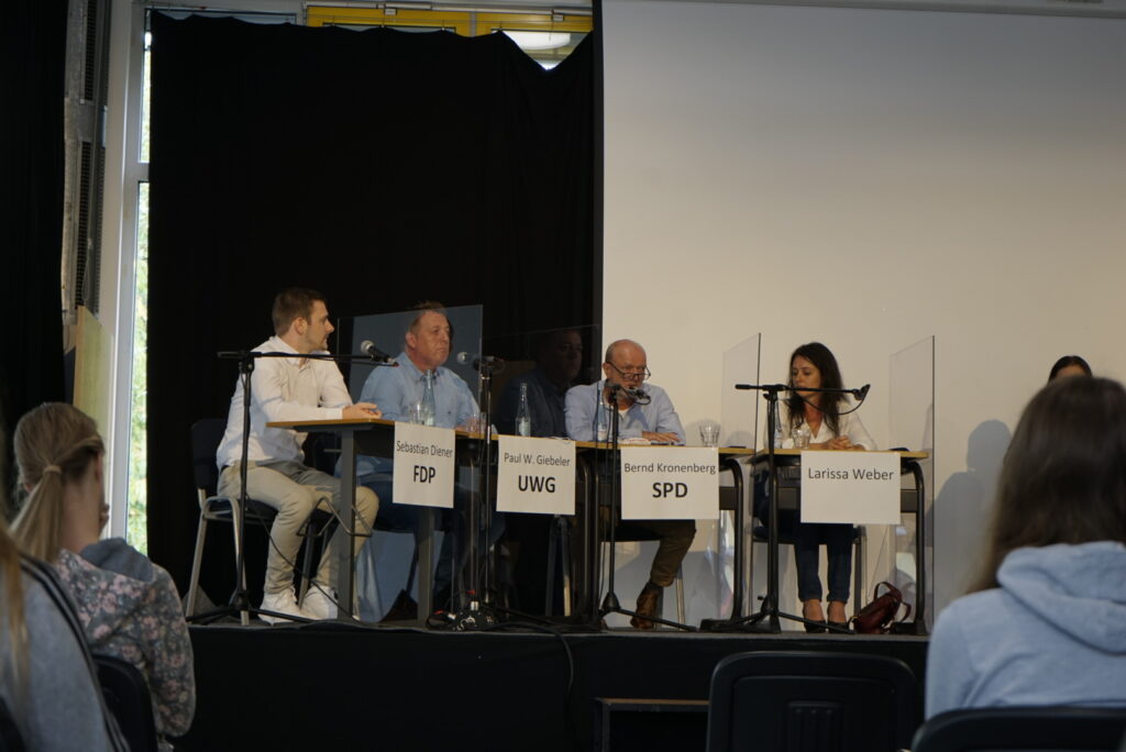 Podiumsdiskussion mit fünf Sprechern an Tischen und Zuhörern im Hintergrund.
