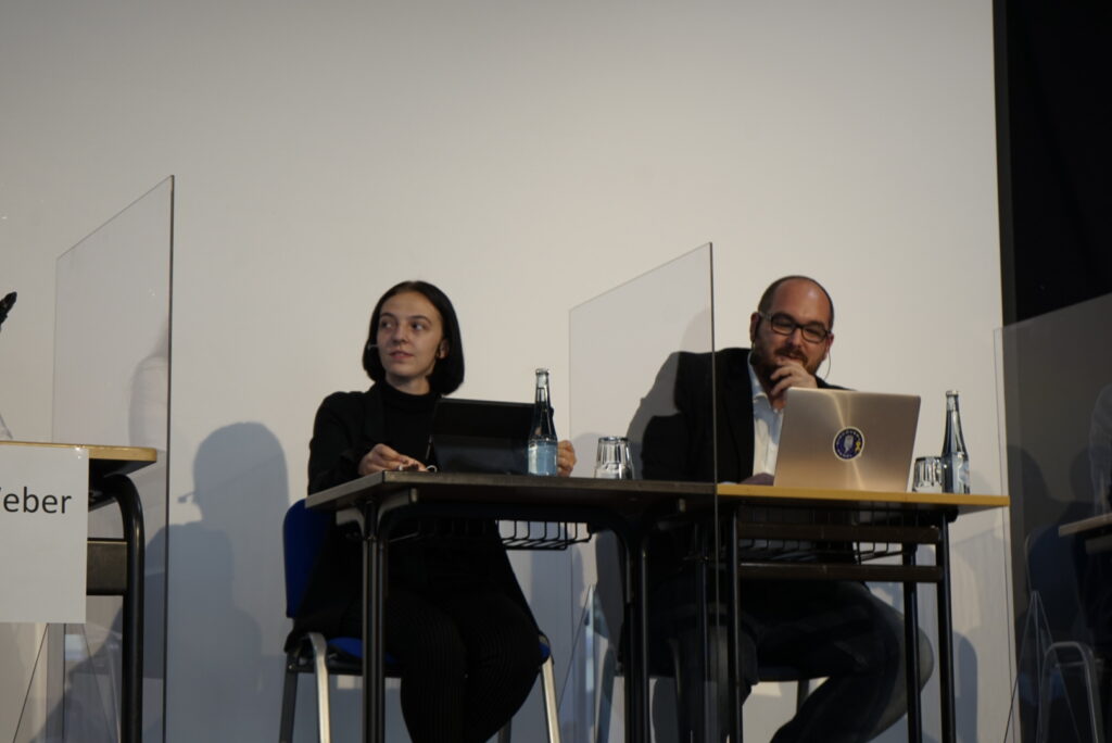 Zwei Personen an einem Tisch mit Laptops und Wasserflaschen, hinter Sichtschutzwänden.