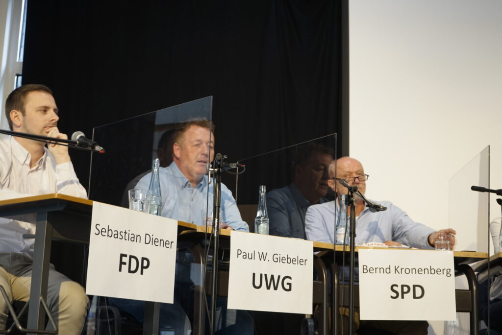 Drei Männer sitzen an einem Tisch bei einer politischen Diskussion mit Mikrofonen.
