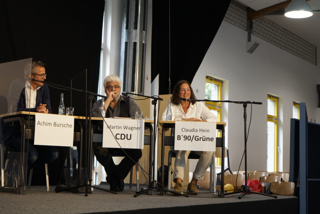 Podiumsdiskussion mit drei Rednern an Tischen und Mikrofonen.