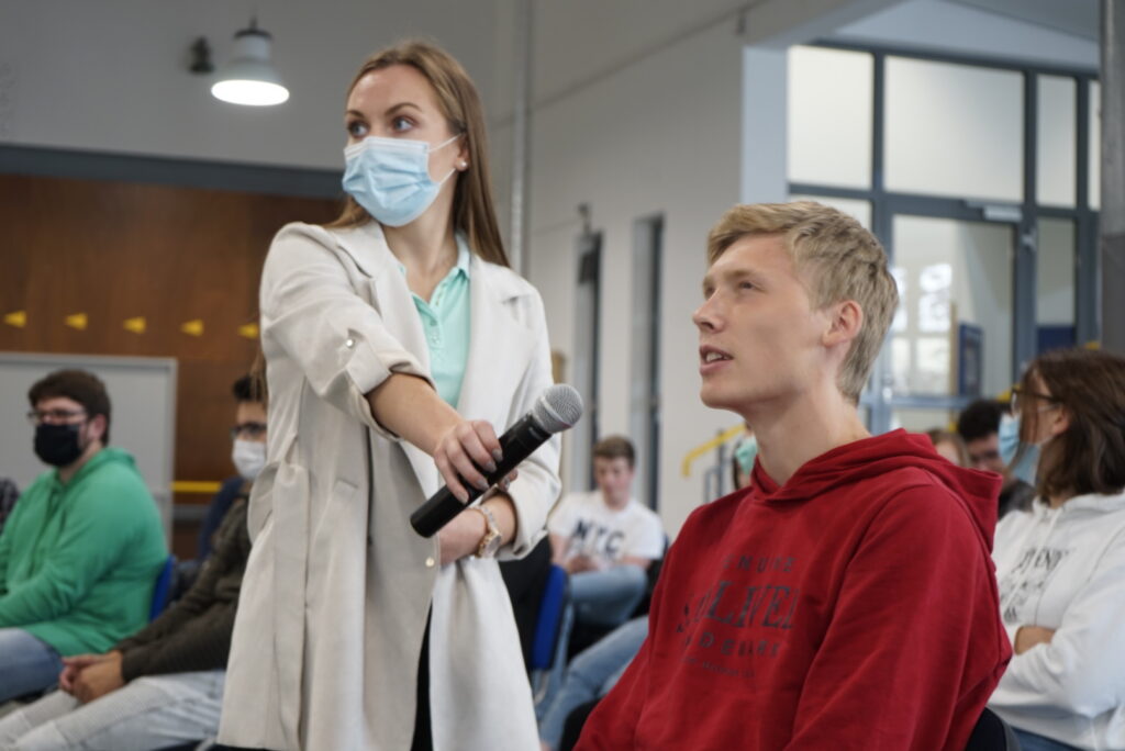 Eine Frau mit Maske hält ein Mikrofon und spricht zu einem Schüler in einem Klassenzimmer.