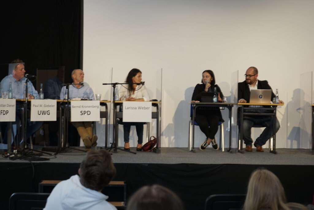 Podiumsdiskussion mit fünf Rednern und moderierender Person auf einer Bühne.