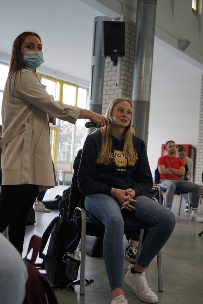 Eine Person spricht mit einem Mikrofon, während eine andere im Sitzen zuhört.