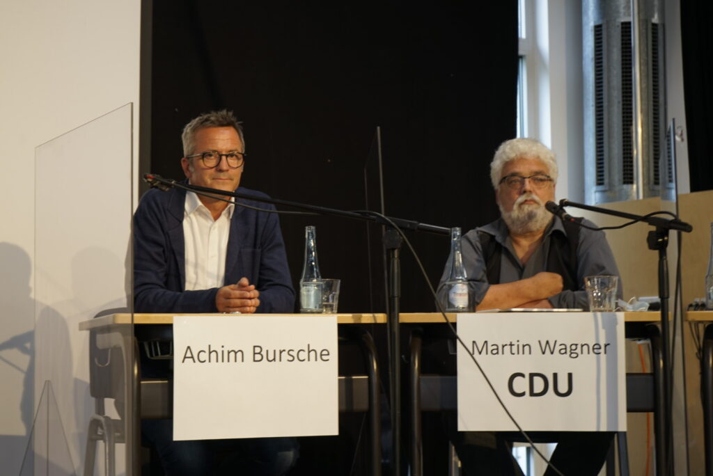 Zwei Männer sitzen an einem Tisch mit Mikrofonen, während einer spricht.