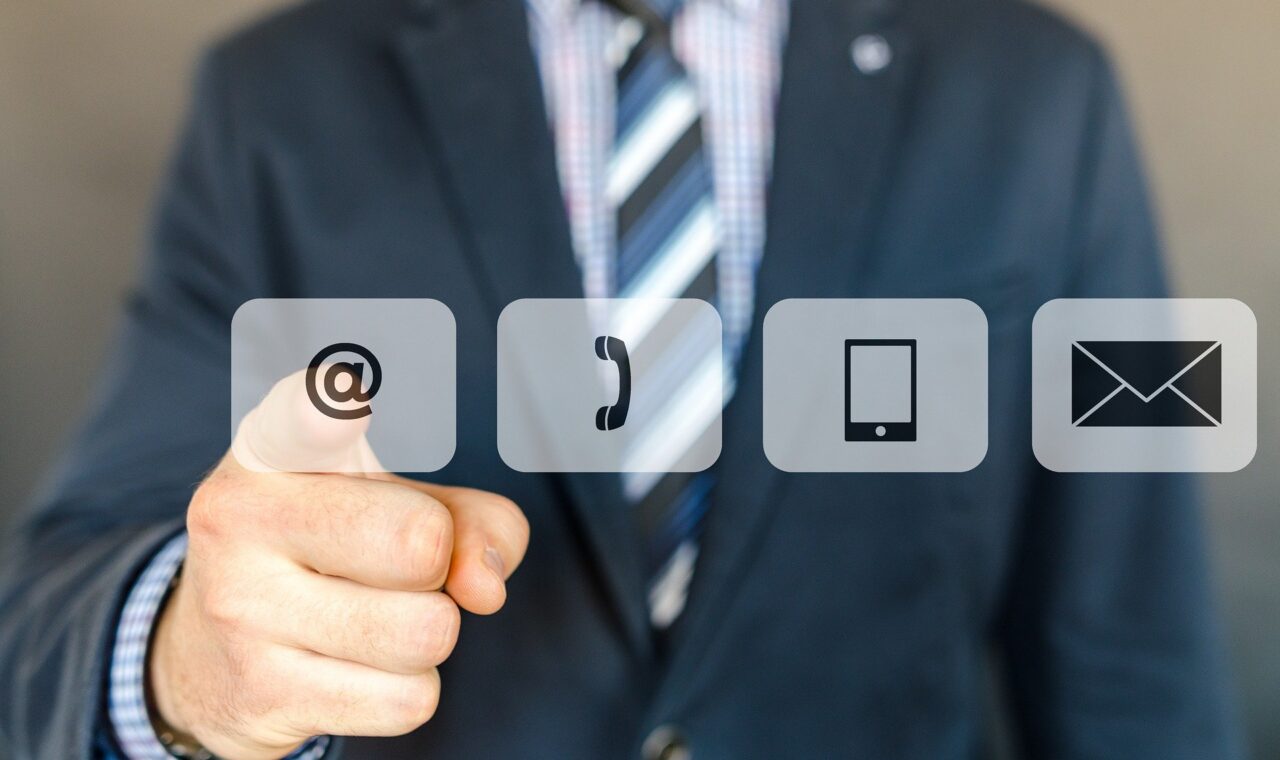 Mann in Anzug zeigt auf Kommunikationssymbole: E-Mail, Telefon, Smartphone.