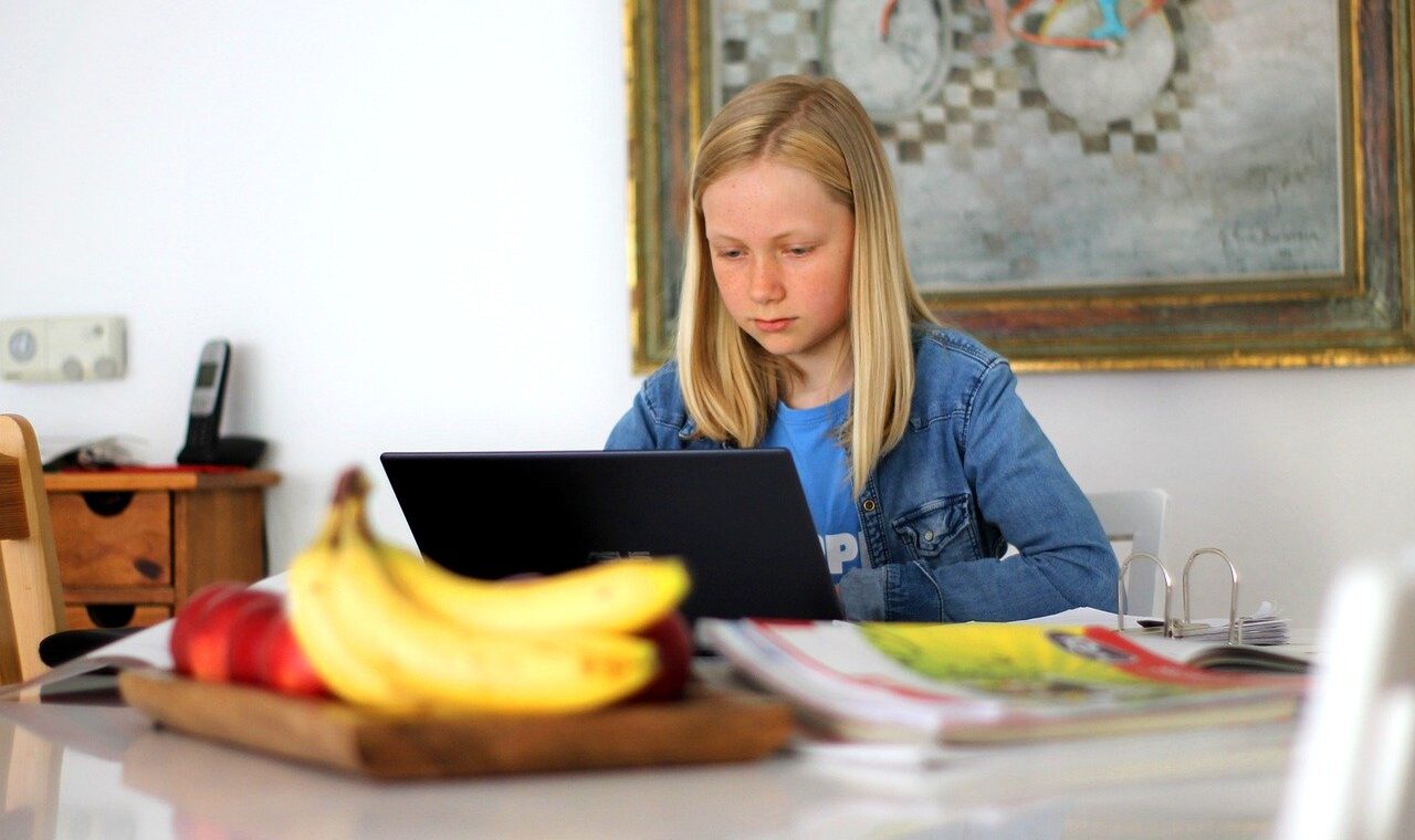 Mädchen sitzt am Tisch und arbeitet am Laptop, im Vordergrund Früchte und Zeitschriften.