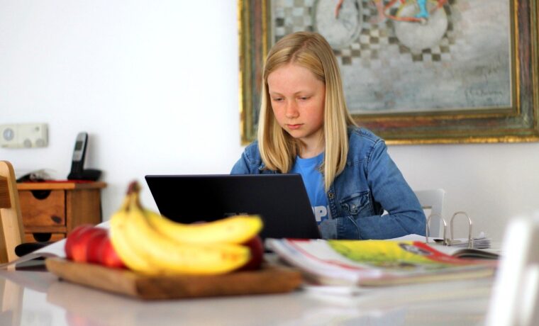 Mädchen sitzt am Tisch und arbeitet am Laptop, im Vordergrund Früchte und Zeitschriften.