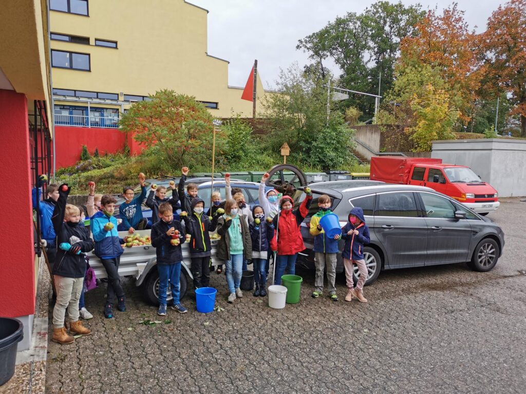 Gruppe von Kindern mit Obst und Gemüse vor einem Auto auf dem Schulgelände.