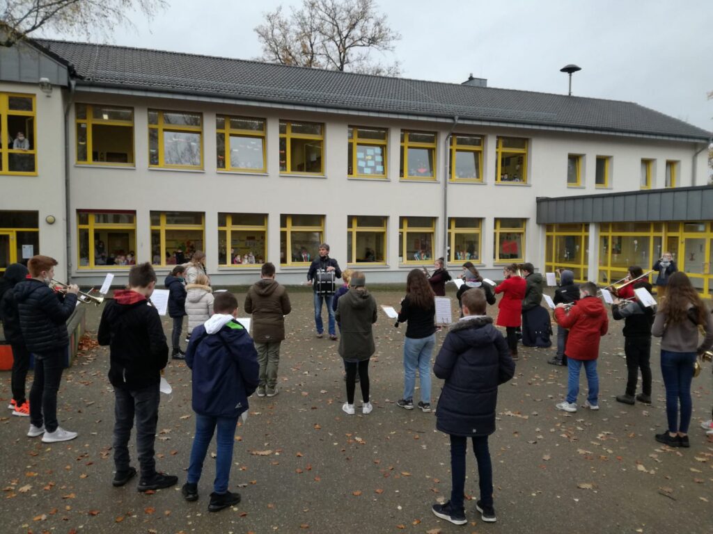 Schüler stehen im Freien und proben mit Notenblättern. Schule im Hintergrund.