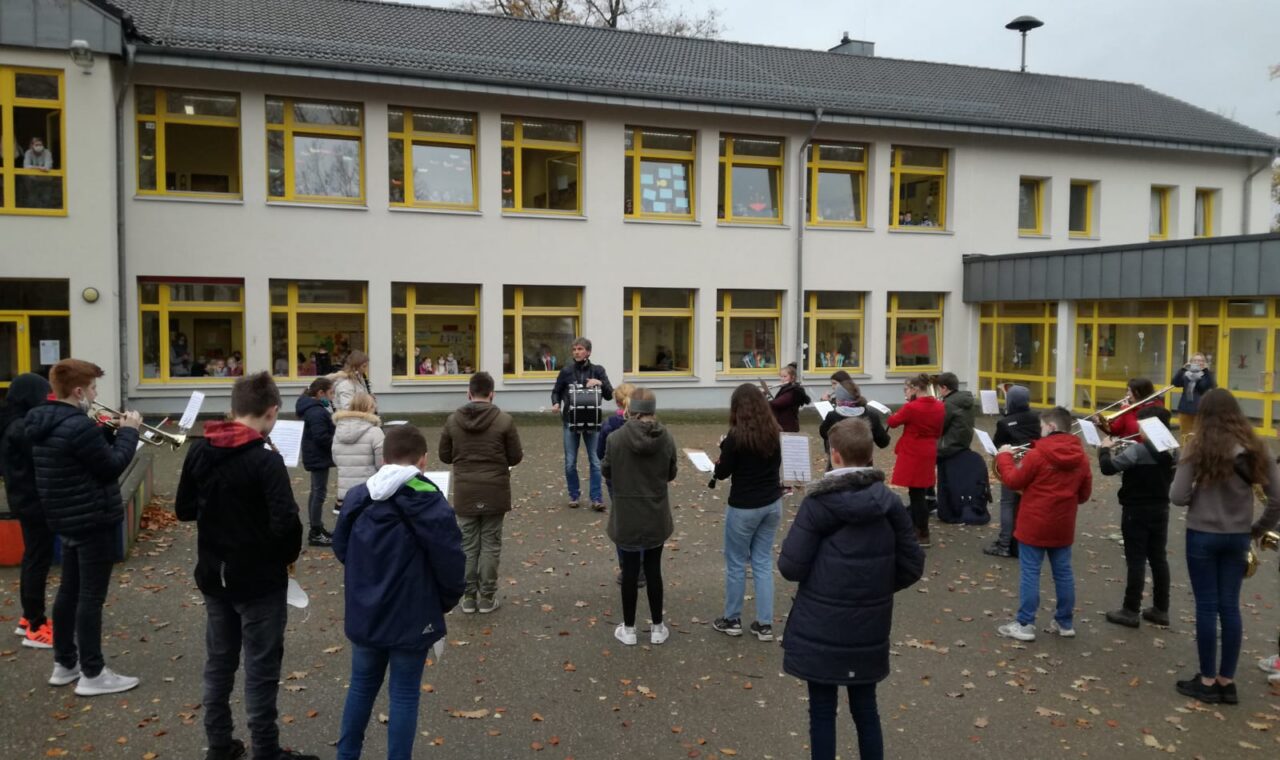 Schüler stehen im Freien und proben mit Notenblättern. Schule im Hintergrund.