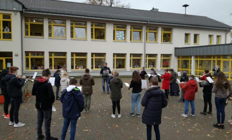 Schüler stehen im Freien und proben mit Notenblättern. Schule im Hintergrund.