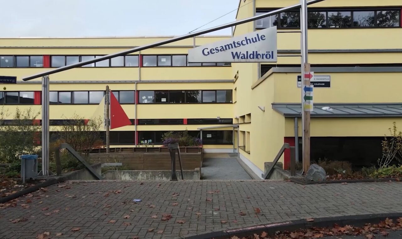 Gesamtschule Waldiefeld, gelbes Gebäude mit Fahnen und Zugang über eine Rampe.