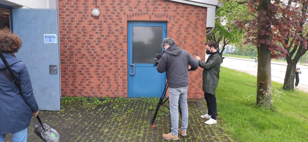 Drei Personen filmen vor einer blauen Tür an einer roten Ziegelfassade.