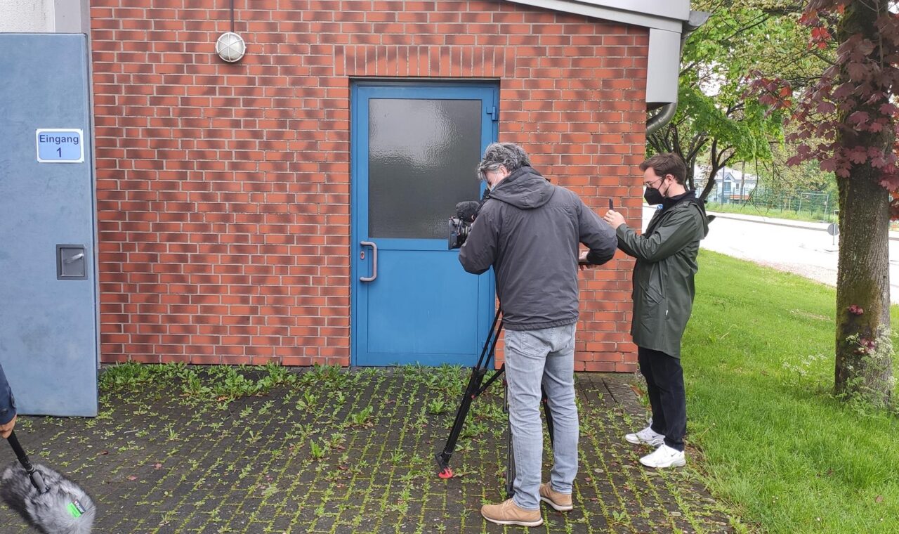 Drei Personen filmen vor einer blauen Tür an einer roten Ziegelfassade.