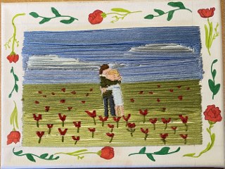 Zwei Personen umarmen sich in einem Feld mit roten Blumen vor blauem Himmel.