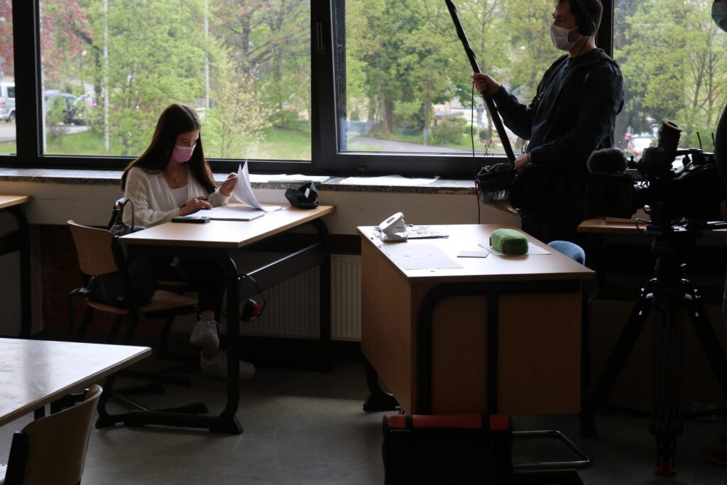 Schülerin arbeitet an ihrem Schreibtisch, Kamerateam im Hintergrund.
