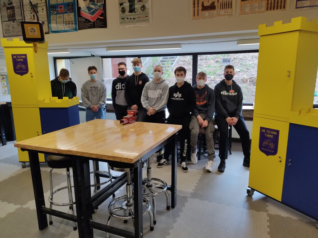 Schüler stehen in einem Klassenraum vor einem Tisch und zwei gelben Schränken.