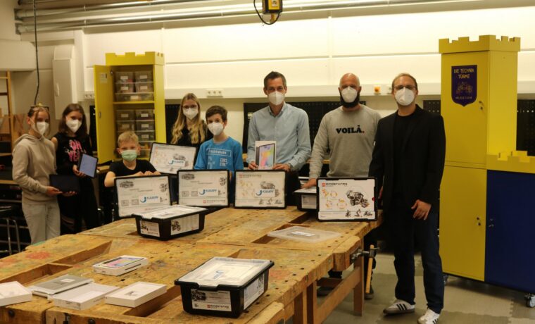 Gruppe von Personen mit Masken präsentiert Projekte in einem Werkraum.