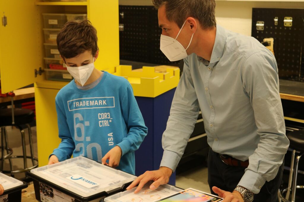 Lehrer zeigt Schülern Materialien am Tisch in einem Klassenraum. Beide tragenMasken.