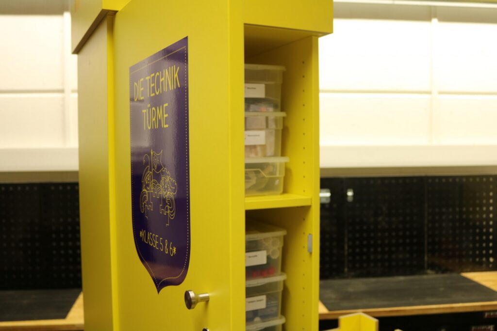 Gelber Schrank mit Fächern und Beschriftungen in einem Klassenraum.