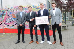 Vier junge Männer in Anzügen stehen vor einer bunten Graffiti-Wand und halten ein Schild.