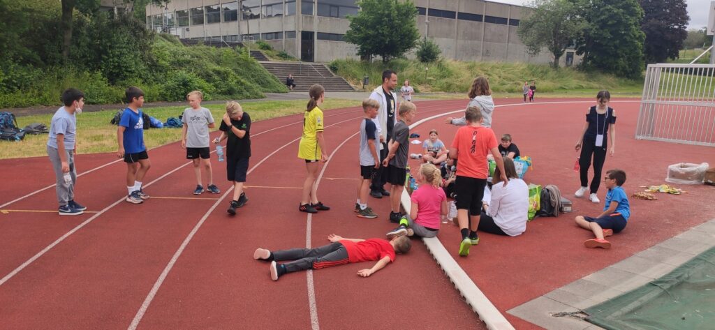 Gruppenspiele auf einem Sportplatz, Kinder in Sportkleidung, einige liegen und ruhen.