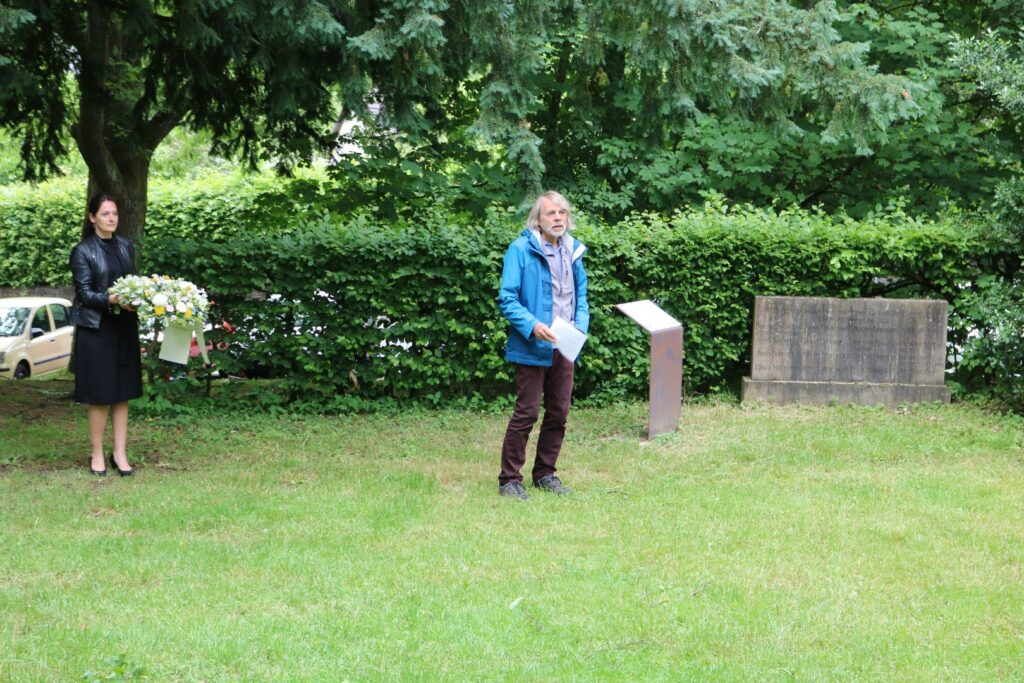 Zwei Personen stehen auf einer Wiese, eine mit Blumen, die andere spricht.