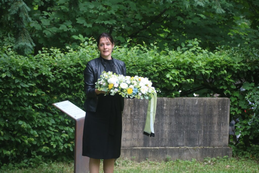 Frau mit Blumenkranz steht vor einer Gedenktafel in der Natur.