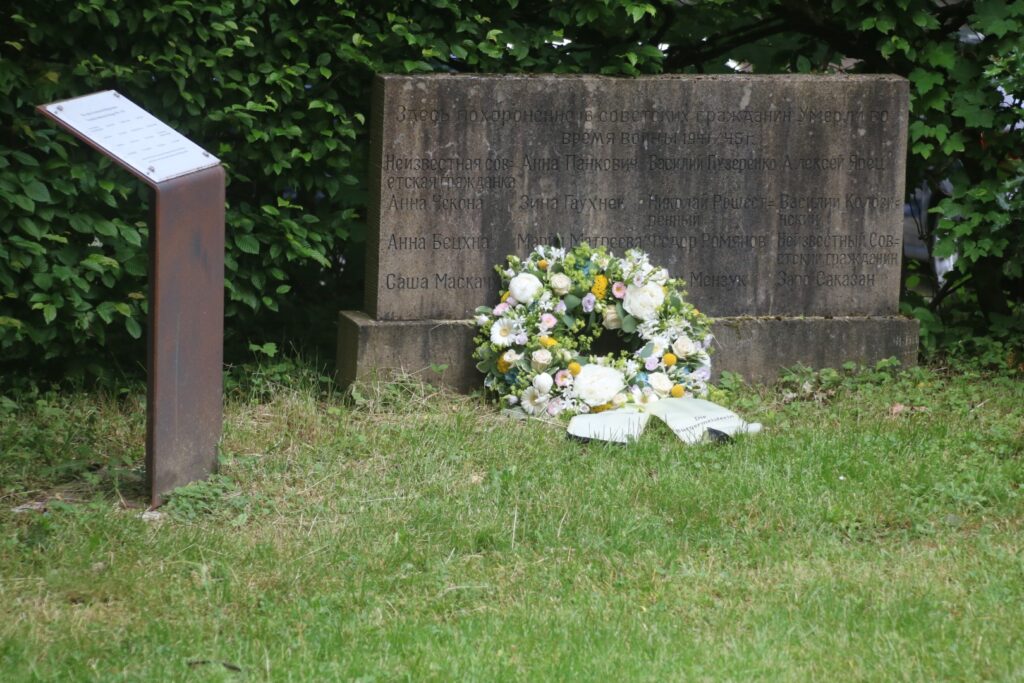 Grabstein mit einem Kranz aus weißen Blumen und einer beschrifteten Tafel daneben.