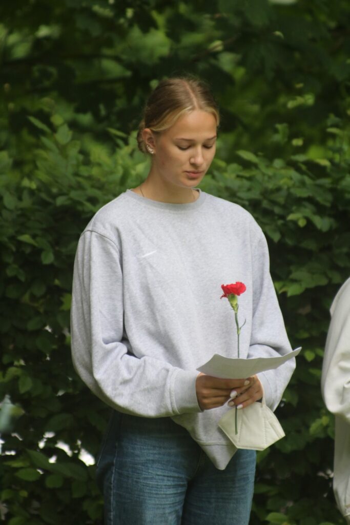 Mädchen hält eine rote Blume und steht vor einer grünen Hecke.