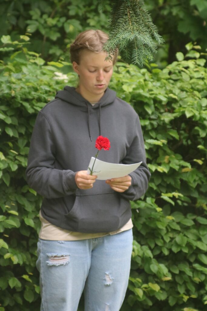 Junge Person hält ein Papier und eine rote Blume vor einer grünen Hecke.