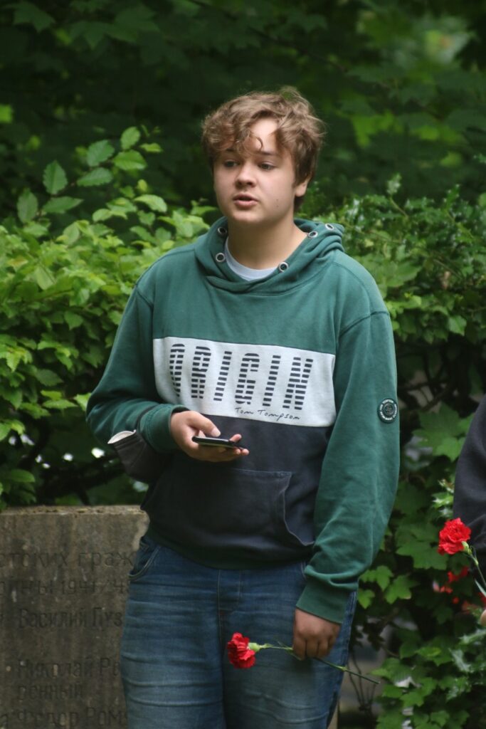 Junger Mann mit Handy und roten Blumen in der Hand, vor grüner Vegetation.