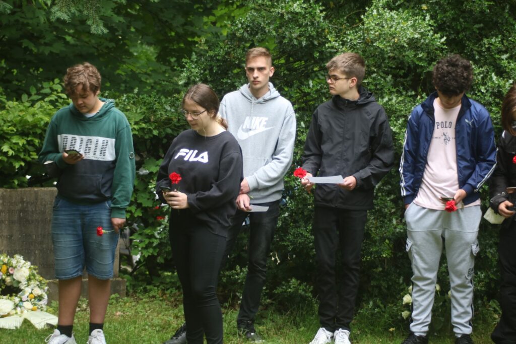 Sechs Jugendliche stehen am Grab und halten Rosen in der Hand.