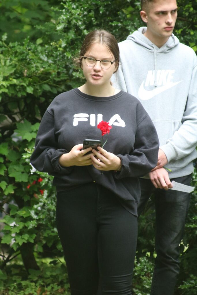 Person mit Smartphone und Rose, im Hintergrund eine weitere Person, grüne Umgebung.