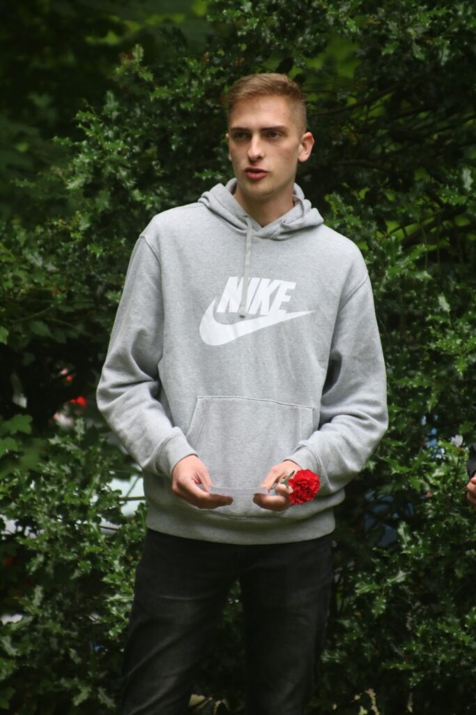 Junger Mann in grauem Hoodie steht vor grünen Pflanzen und hält eine rote Blume.