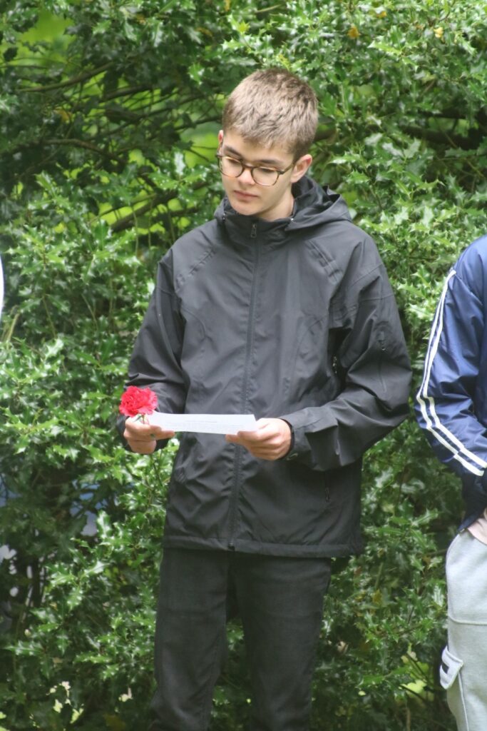 Jugendlicher in schwarzer Jacke hält einen Zettel und eine rote Blume.